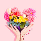 Colorful flowers arranged in a heart shape. Two hands are framing the base of the flower heart. This is in front of a light pink backdrop