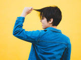 A girl with brown pixie cut hair is standing in front of an orange wall with her back to us She is pulling her bangs up with one hand and wearing a denim jacket.