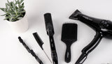 black hairdryer, hairbrush, flat iron, comb, and curling wand laid out next to a potted plant.